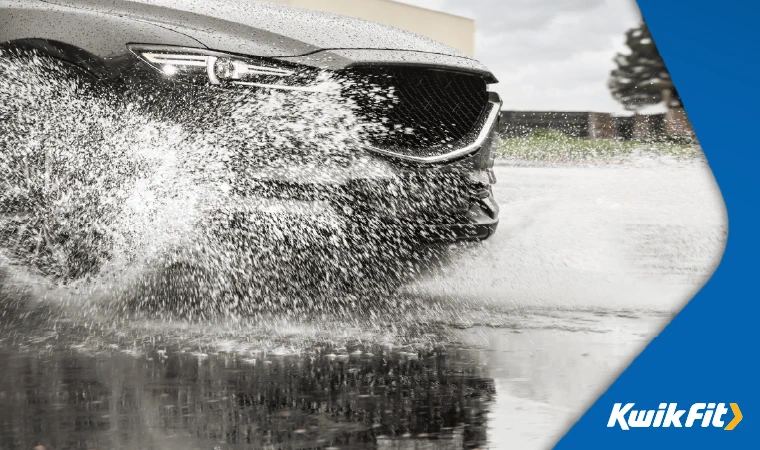 Grey car driving through a big puddle with water splashing up on to the car.